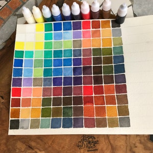 May include: A watercolor paint chart with 64 color swatches arranged in a triangular pattern. The colors range from yellow to red to blue to green to brown. The chart is labeled "Jazzer Stardust Handmade".