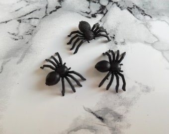 May include: Three small, black plastic spiders with eight legs each are arranged on a white and gray marbled surface. The spiders have a rounded body and are a common Halloween decoration.