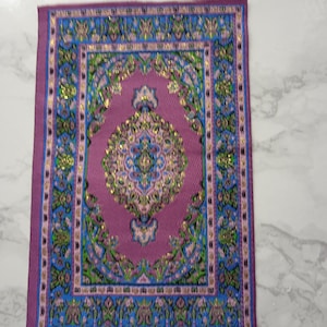 Puede incluir: Una alfombra rectangular en miniatura con un diseño de medallón central en tonos morado, azul, verde y dorado. La alfombra presenta un borde ornamentado y patrones intrincados, mostrando una estética tradicional. La alfombra es probablemente para una casa de muñecas o un entorno en miniatura.