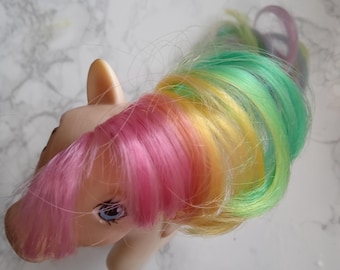 May include: A vintage toy pony with a cream-coloured body and a rainbow-coloured mane and tail. The mane features pink, yellow, and green strands. The pony has blue eyes and is lying on a white marble surface.