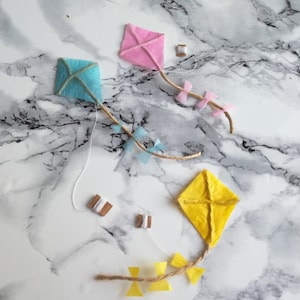 May include: Three paper kites, one blue, one pink, and one yellow, are laid out on a white marble surface. Each kite has a string attached to it, and there are small spools of string scattered around the kites.