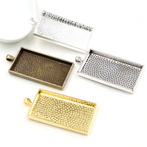 May include: Four rectangular pendant trays in silver, bronze, and gold tones. Each tray has a textured surface and a loop for attaching to a chain or cord. The trays are empty, ready for use in jewellery making or crafting projects.