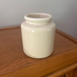 May include: A small, cylindrical ceramic jar with a smooth, off-white glazed finish. The vessel features a slightly rounded rim and a straight body that tapers gently toward the base, presenting a clean, simple, and functional design for storage.