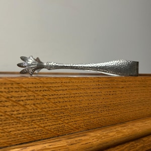 May include: A silver-toned ice tong with a textured handle and a claw-like grip. The tongs are made of metal and have a detailed, hammered design. The claw-shaped end adds a unique, decorative element to the barware.