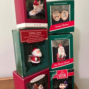 May include: A collection of Hallmark Keepsake Christmas ornaments in their original boxes. The boxes are red and green, featuring various holiday-themed figurines, including Santa Claus, gnomes, and woodland creatures. The boxes are labeled with the ornament names.