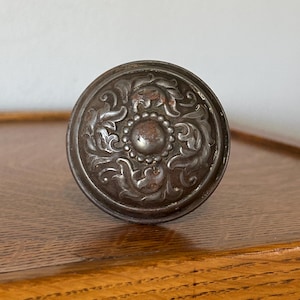 May include: Antique silver-toned cabinet knob with an ornate floral design. The knob is round with a central, raised circular element surrounded by detailed, swirling patterns. The metal has a slightly aged appearance, suggesting vintage craftsmanship.