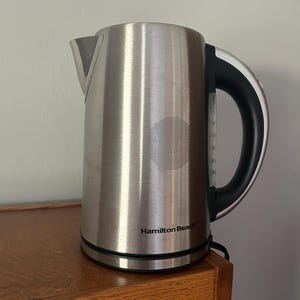 May include: Stainless steel Hamilton Beach electric kettle. The kettle has a black handle and a spout. The brand name is printed on the front. The kettle is sitting on a wooden surface.