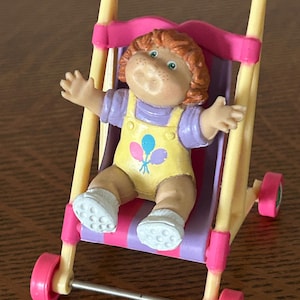 May include: A vintage Cabbage Patch Kids doll in a yellow and purple outfit, seated in a pink and yellow toy stroller. The doll has red hair, freckles, and is smiling. The stroller has pink wheels.