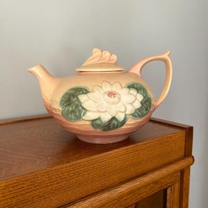 May include: A peach-colored ceramic teapot with a floral design. The teapot features a white flower with a red center and green leaves. The teapot is sitting on a wooden surface.