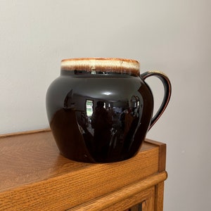 May include: A brown glazed ceramic pitcher with a white rim and a brown handle. The pitcher is sitting on a wooden surface.