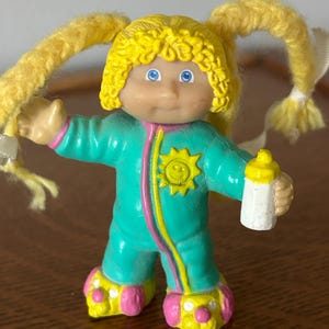 May include: A vintage doll figurine with yellow curly hair in pigtails, blue eyes, and a smiling face. The doll wears a teal onesie with a sun graphic and pink and yellow accents, holding a white bottle with a yellow cap.