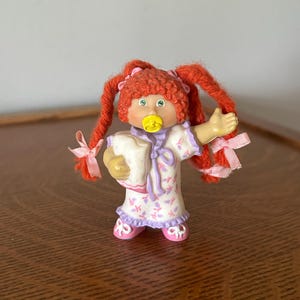 May include: A vintage doll with red yarn pigtails, wearing a white and lavender floral print robe and pink shoes. The doll has a pacifier and is holding a small white pillow. The doll is a collectible toy.