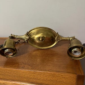 May include: Antique brass light fixture with two downward-facing light sockets. The fixture has an oval central plate and ornate, decorative arms. The overall design is vintage, with a warm, golden hue.