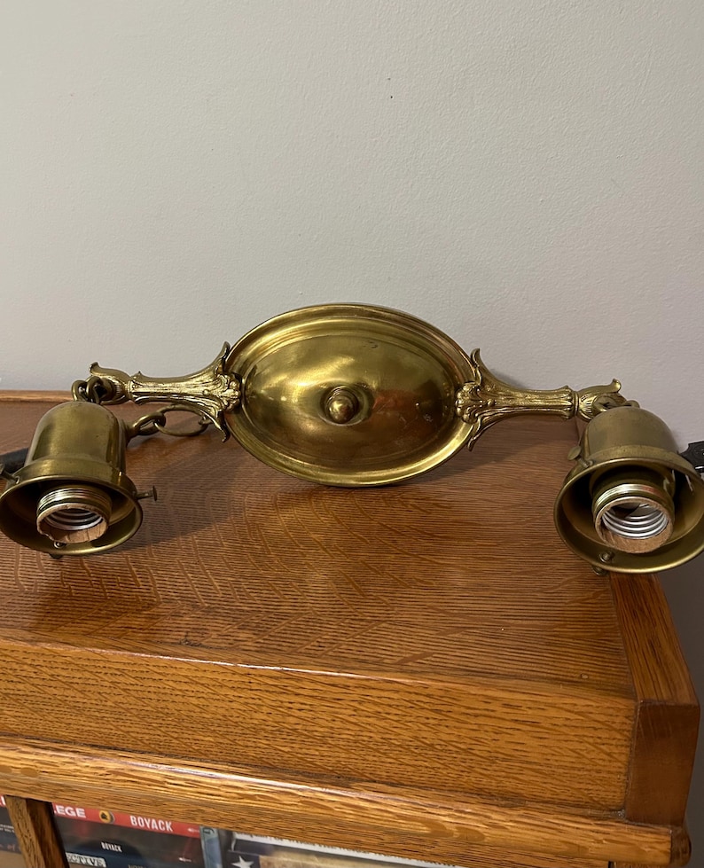 May include: Antique brass light fixture with two downward-facing light sockets. The fixture has an oval central plate and ornate, decorative arms. The overall design is vintage, with a warm, golden hue.