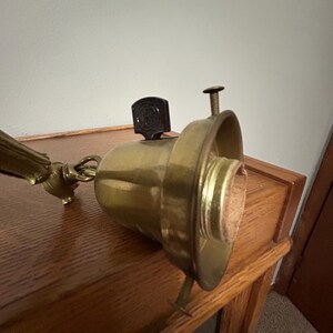 May include: Vintage brass lamp fixture with a dark brown switch and a screw. The fixture has a gold finish and is mounted on a wooden surface. The lamp is designed to hold a light bulb.