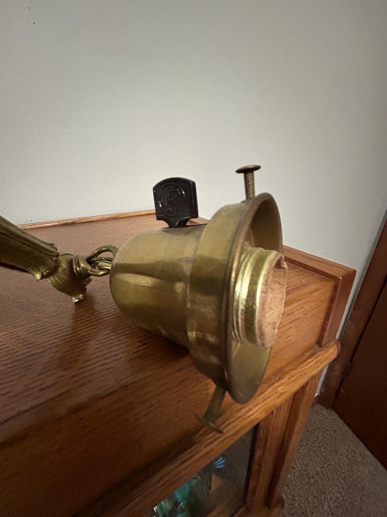 May include: Vintage brass lamp fixture with a dark brown switch and a screw. The fixture has a gold finish and is mounted on a wooden surface. The lamp is designed to hold a light bulb.