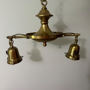 May include: A vintage brass chandelier with two bell-shaped light fixtures. The chandelier is suspended from a chain and features a central decorative element. The brass has a warm, aged patina, suggesting its antique nature. The design is classic and elegant.