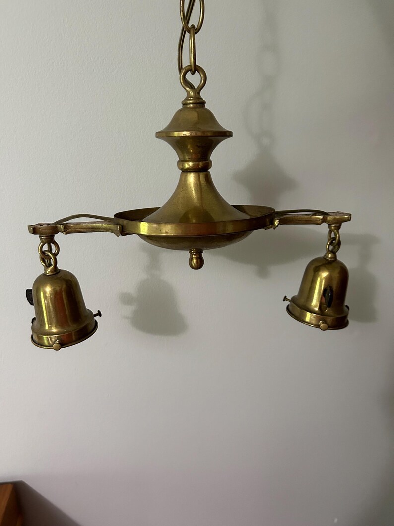 May include: A vintage brass chandelier with two bell-shaped light fixtures. The chandelier is suspended from a chain and features a central decorative element. The brass has a warm, aged patina, suggesting its antique nature. The design is classic and elegant.