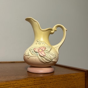 May include: A small, vintage-style pitcher with a light yellow body and a pink base. The pitcher features a floral design with pink and green accents. It has a curved handle and a spout for pouring. The pitcher is likely made of ceramic.