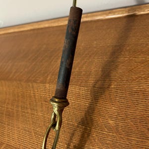 May include: Close-up of a vintage lamp fixture. The fixture features a dark, cylindrical metal rod with visible rust, connected to a brass-colored chain. The chain is linked to a brass-colored fitting. The background is a wooden surface.