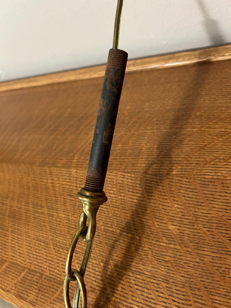 May include: Close-up of a vintage lamp fixture. The fixture features a dark, cylindrical metal rod with visible rust, connected to a brass-colored chain. The chain is linked to a brass-colored fitting. The background is a wooden surface.