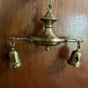 May include: Vintage brass chandelier with two bell-shaped light fixtures. The chandelier is suspended from a chain and features an ornate design. The brass has a warm, aged patina, adding to its antique charm. The light fixtures are open at the bottom.