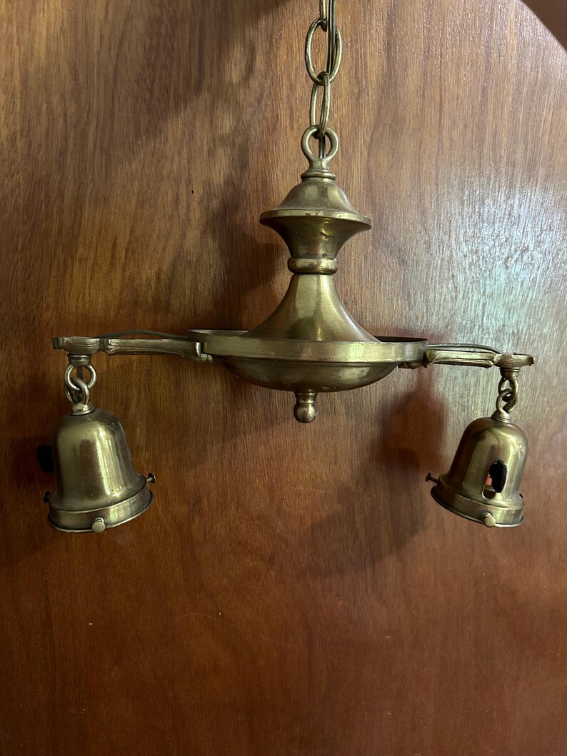 May include: Vintage brass chandelier with two bell-shaped light fixtures. The chandelier is suspended from a chain and features an ornate design. The brass has a warm, aged patina, adding to its antique charm. The light fixtures are open at the bottom.