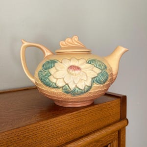 May include: A ceramic teapot with a floral design. The teapot is peach-colored with a large white flower and green leaves on the front. The lid and handle are also peach-colored. The teapot is sitting on a wooden surface.