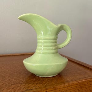May include: A light green ceramic pitcher featuring a wide, rounded base that tapers into a narrow neck with three horizontal ribbed bands. The pitcher has a curved handle on the side and a prominent, elongated pouring spout at the top rim.