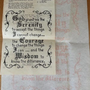 May include: A cream-colored fabric paint-by-number sampler featuring the Serenity Prayer in black decorative lettering, surrounded by ornate scrollwork borders. The sheet includes printed instructions and a color key, with a secondary translucent transfer sheet showing the same design in red outlines.