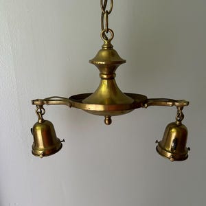 May include: A vintage brass chandelier with two bell-shaped light fixtures. The chandelier is suspended from a chain and features a central decorative element. The brass has a warm, aged patina, suggesting it is an antique or vintage piece.