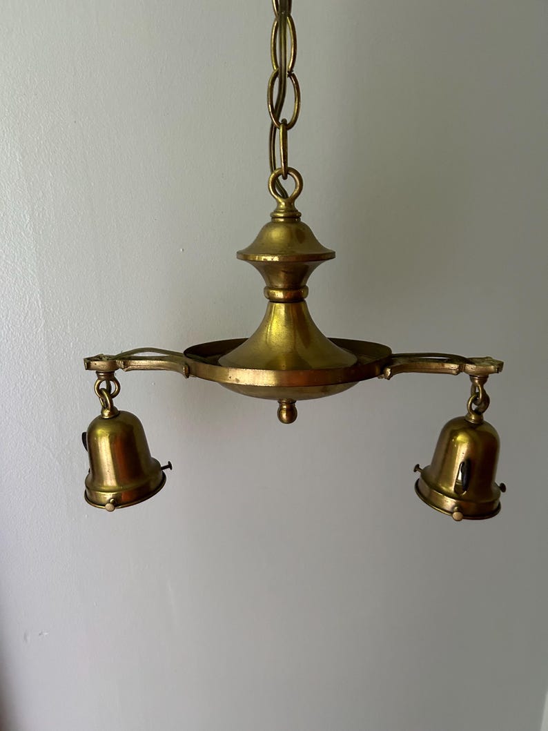 May include: A vintage brass chandelier with two bell-shaped light fixtures. The chandelier is suspended from a chain and features a central decorative element. The brass has a warm, aged patina, suggesting it is an antique or vintage piece.