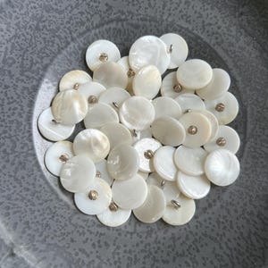 May include: A collection of round, white mother-of-pearl buttons with gold-colored metal shanks. The buttons are arranged in a gray bowl, showcasing their iridescent surfaces and classic design. Ideal for sewing and craft projects.