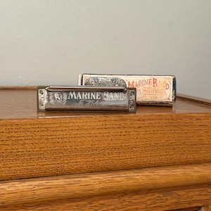 May include: A silver Marine Band harmonica with the text "MARINE BAND" and a decorative emblem. The harmonica rests on a wooden surface next to its original box, which features the text "MARINE B D A. HOHNER".