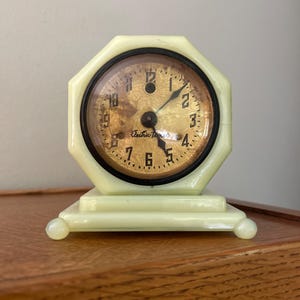May include: An octagonal pale yellow clock with a round face featuring a textured gold background, black numerals, and black hands. The clock face displays the text Electric Time. It sits on a rectangular base with four small rounded feet.