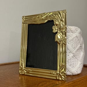 May include: A rectangular gold-colored picture frame with a polished metallic finish. The frame features raised, molded floral and leaf-like decorative accents at the corners and along the edges. It contains a solid black insert panel.