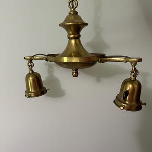 May include: A vintage brass chandelier with two bell-shaped light fixtures. The chandelier has a central decorative element and is suspended from a chain. The brass has a warm, aged patina, suggesting it is an antique.