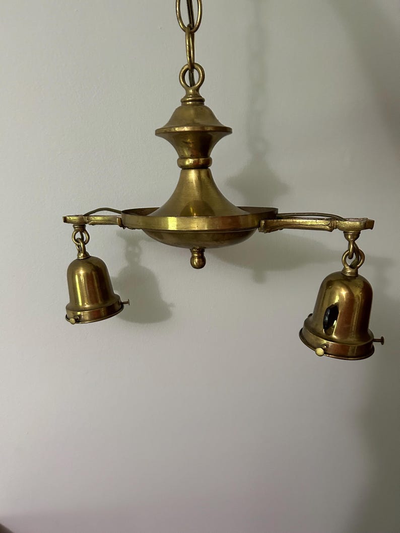 May include: A vintage brass chandelier with two bell-shaped light fixtures. The chandelier has a central decorative element and is suspended from a chain. The brass has a warm, aged patina, suggesting it is an antique.