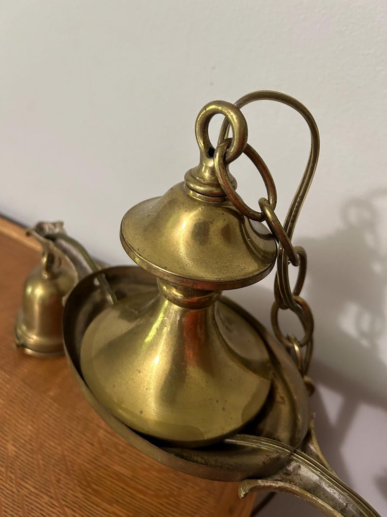 May include: Close-up of a vintage brass chandelier fixture. The fixture features a decorative bowl, a central stem, and a chain for hanging. The brass has a warm, aged patina, suggesting its antique nature. The design is ornate and detailed.