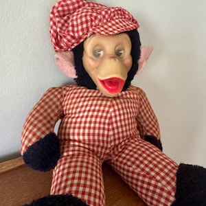 May include: A vintage monkey doll with a red and white checkered outfit and matching cap. The doll has a tan face, red lips, and black fur around the face and limbs. The cap is topped with a blue pom-pom. The doll is sitting on a wooden surface.