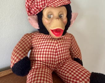 May include: A vintage monkey doll with a red and white checkered outfit and matching cap. The doll has a tan face, red lips, and black fur around the face and limbs. The cap is topped with a blue pom-pom. The doll is sitting on a wooden surface.