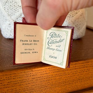 May include: A small red leather-bound book with the title "Petite Calendar and Stamp Case" and the year "1908" printed on the front cover. The book is open to reveal a page with the text "Compliments of Frank Le Bron Jewelry Co. 404 Main St. Keokuk, Iowa."