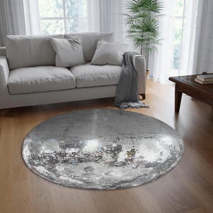 Può includere: Un tappeto rotondo argentato con un design a palla da discoteca a specchio. Il tappeto si trova in un soggiorno con un divano e una pianta.