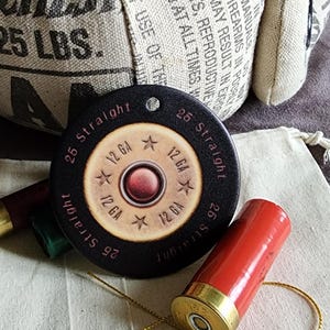 May include: A black, round pendant with the text "25 Straight" and "12 GA" encircling a central design. The pendant is near red and green shotgun shells and a canvas bag. The background includes a burlap sack with text.