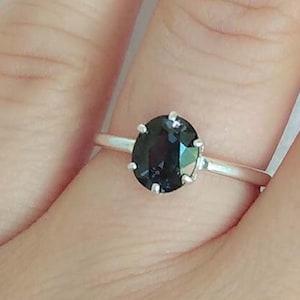 May include: A silver ring featuring an oval-cut, dark gemstone. The gemstone is set in a classic prong setting, and the band is a simple, polished silver. The ring is shown on a person's finger.