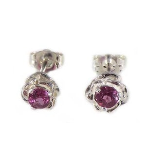 Puede incluir: Un par de pendientes de plata con cierre de tuerca, cada uno con una gema redonda de color rosa intenso engastada en un diseño floral. Los pendientes se presentan sobre un fondo blanco, resaltando su diseño y color.