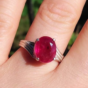 Puede incluir: Un anillo de plata con una gran piedra preciosa de rubí de forma ovalada. El rubí es de color rojo intenso y está engastado en un engaste simple y elegante.