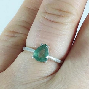 Puede incluir: Anillo de plata con una piedra preciosa verde esmeralda en forma de pera. La banda del anillo es delgada y sencilla, con la piedra engastada en un engaste de cuatro garras. La piedra tiene una superficie pulida.