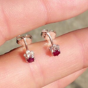 Op de afbeelding: Een paar kleine oorstekers met een zilveren pin en zetting. Elke oorbel heeft een ronde, dieprode edelsteen. De oorbellen worden gepresenteerd tegen een vage achtergrond.