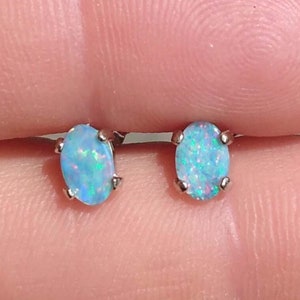 May include: A pair of silver stud earrings with oval-shaped opal stones. The opals are a light blue color with iridescent green and white flecks.